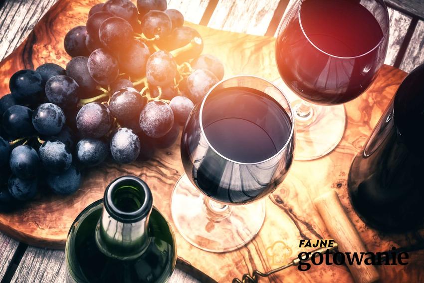 Wino z winogron podane w kieliszkach. Obok nich stoi butelka z winem oraz leżą winogrona.