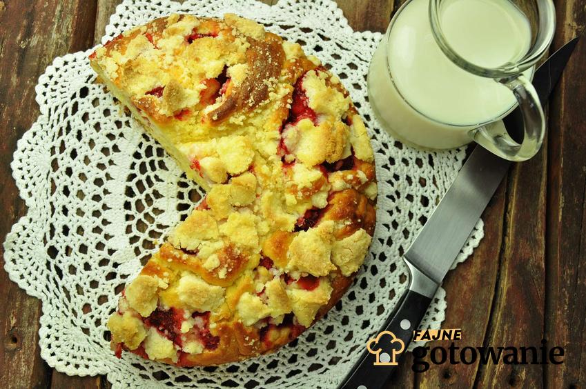 Ciasto drożdżowe z truskawkami podane na szydełkowej serwetce, z dzbankiem mleka i nożem na drewnianym stole.