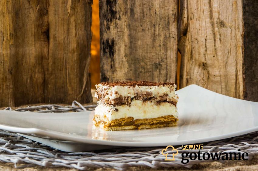 Ciasto bez pieczenia na herbatnikach podane jest na białym, prostokątnym talerzu. W oddali można zauważyć ścianę z drewnianych desek.